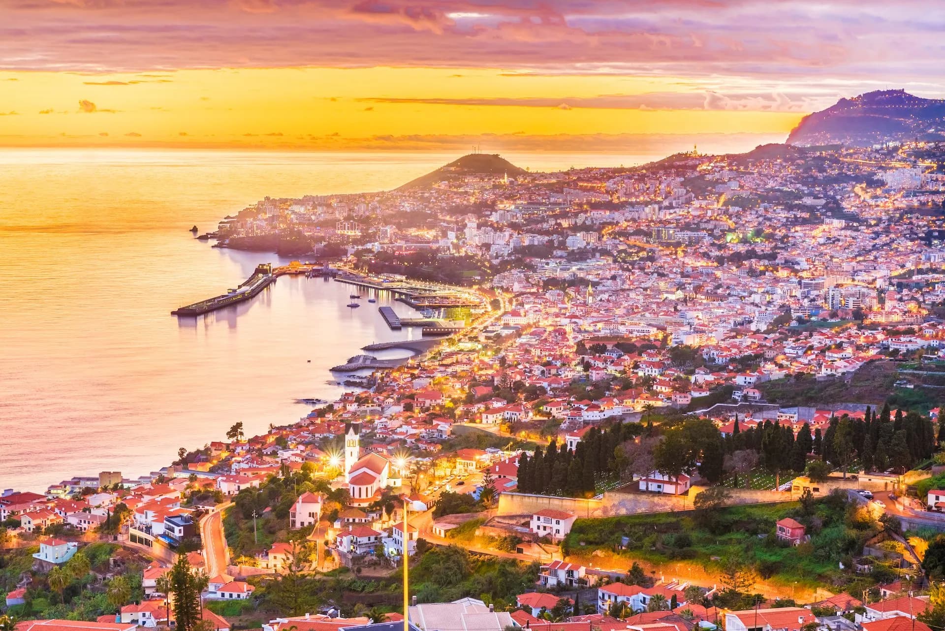 Funchal city lights along the coast at sunset with warm orange and purple sky over the sea.