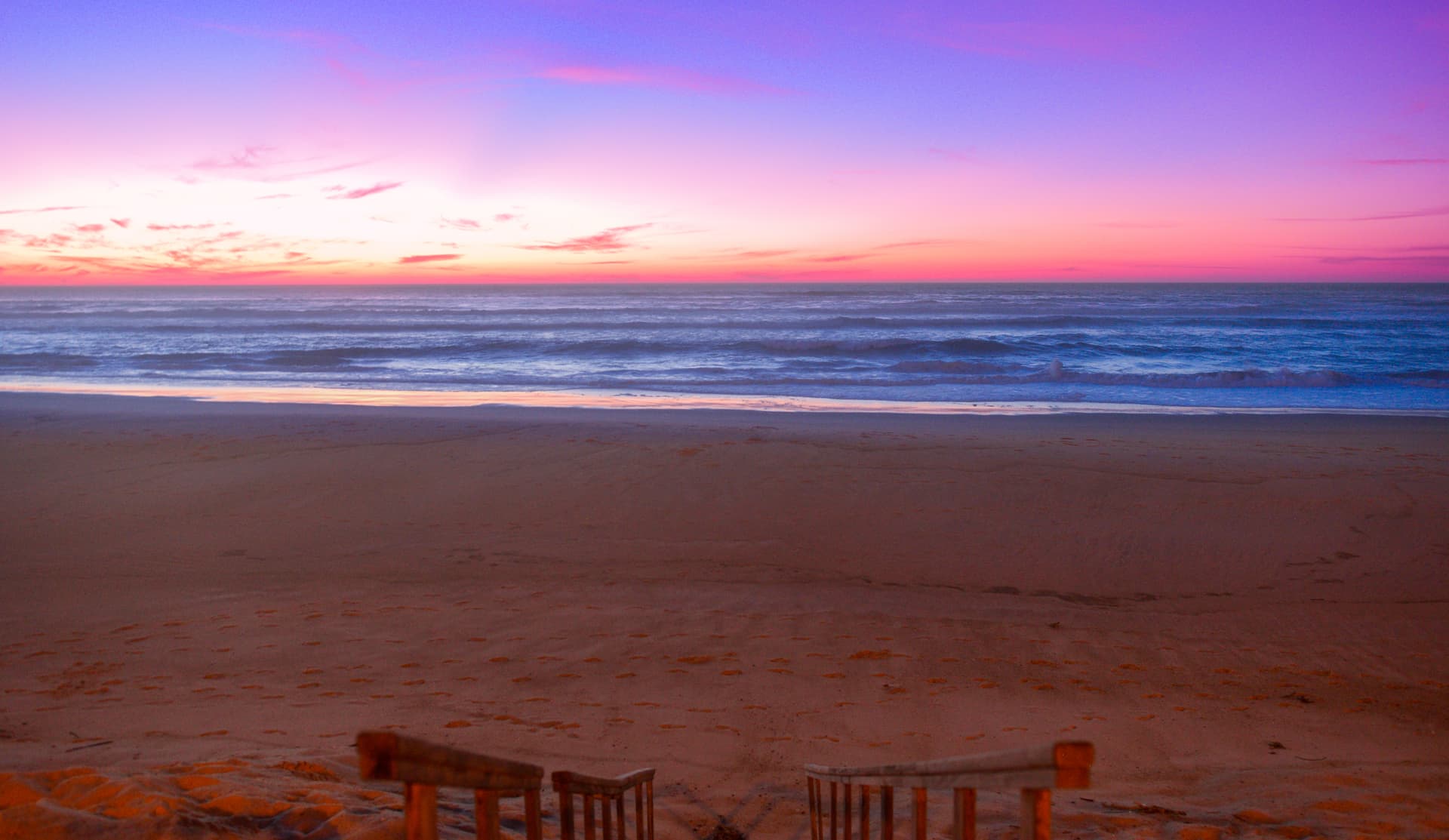 Beach with wooden steps leading down to sand, ocean waves, and a vibrant pink and purple sunset.