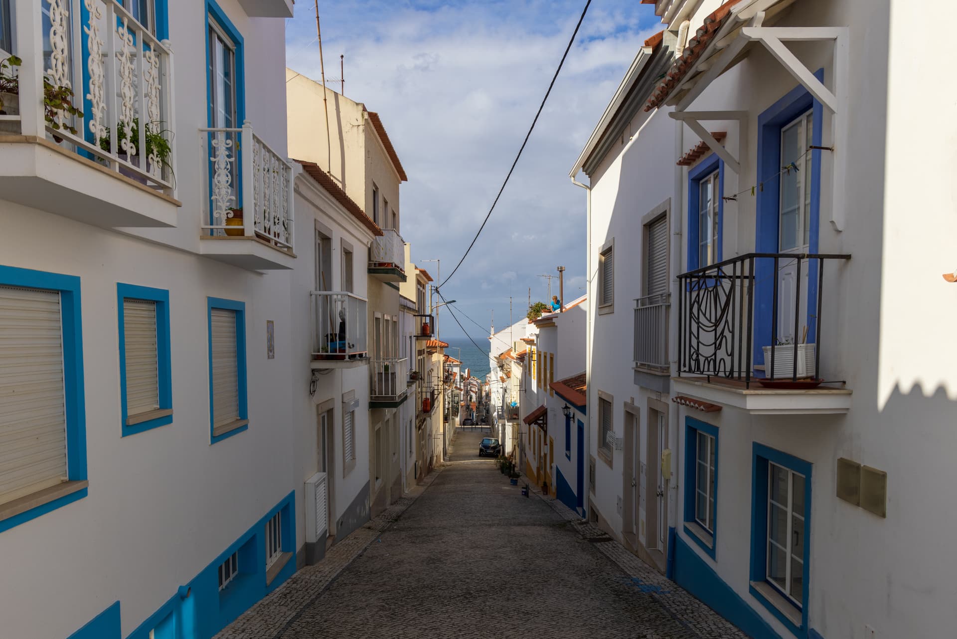 Cobblestone street sloping down between white buildings toward the sea in Nazare.