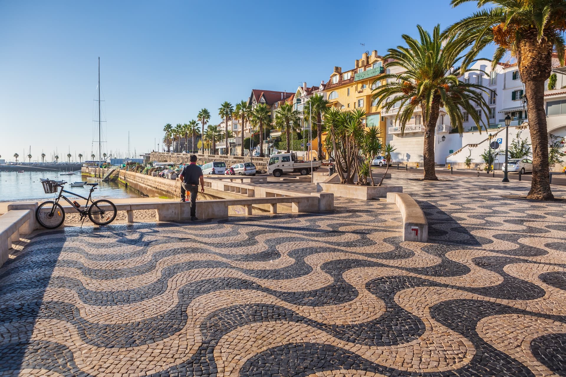 Cobblestone promenade with wave pattern, palm trees, and harbor in Cascais