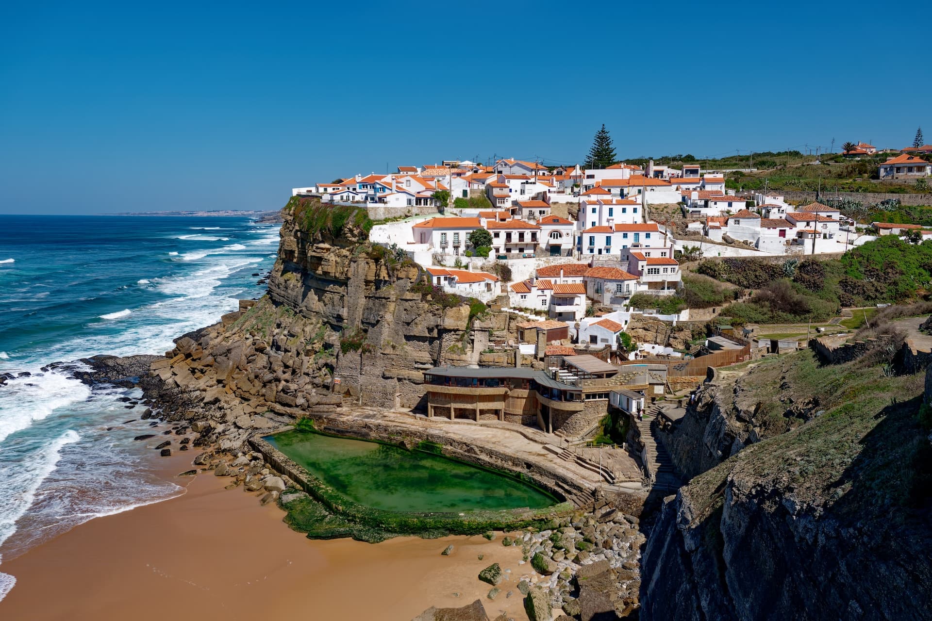 White village houses on cliffside above beach and green tidal pool in Azenhas do Mar.