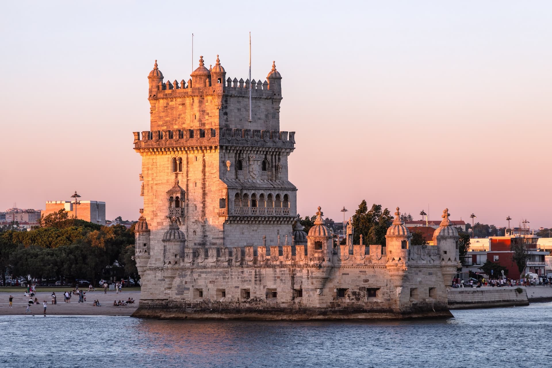 Belém Tower illuminated by sunset light, standing in the water near a crowded shore.