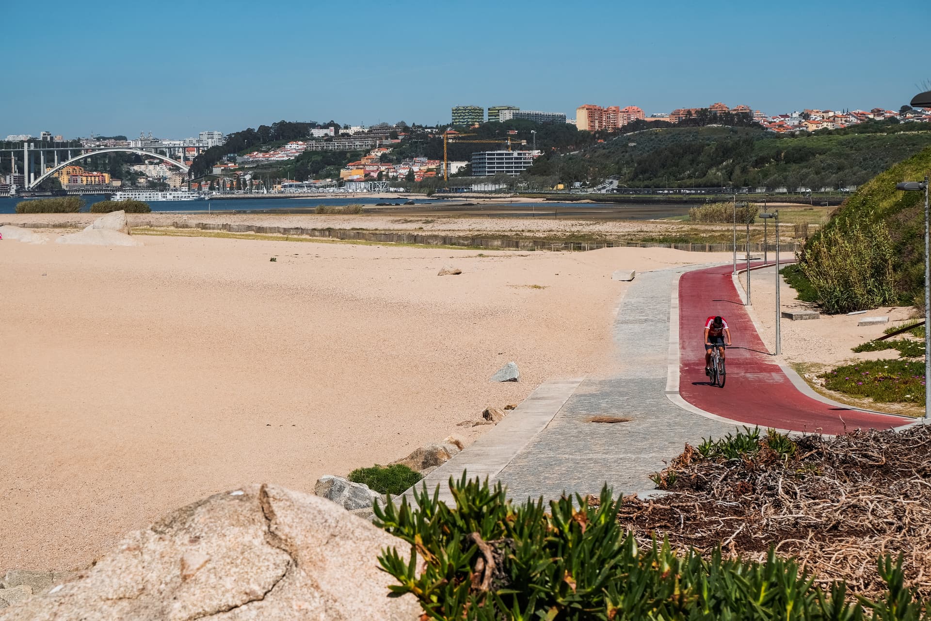 Cyclist on red bike path beside sandy beach with city and bridge in background, Portugal.