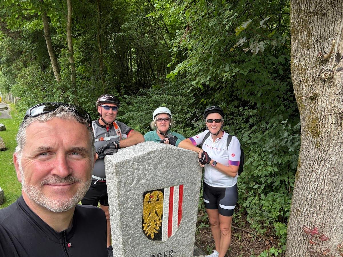 Cyclists taking a selfie by a stone marker with a coat of arms in a wooded area.