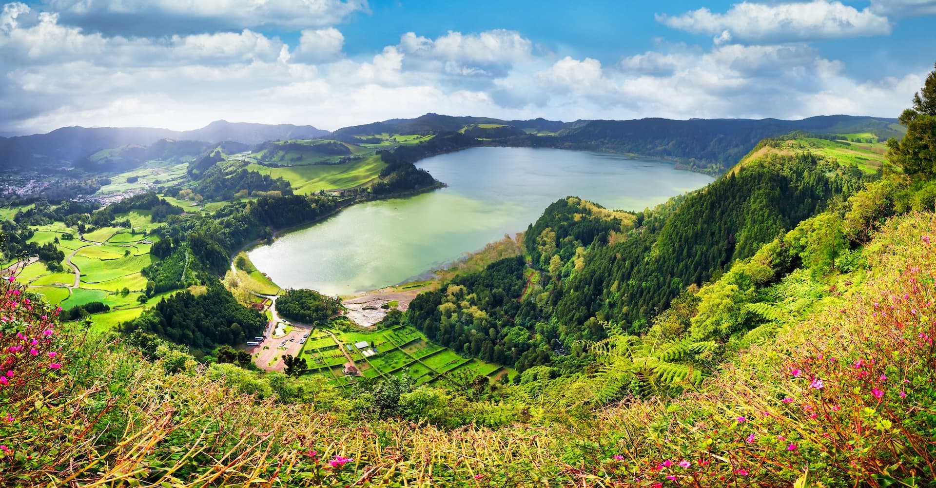 Lagoa das Furnas lake surrounded by green hills, forests, and fields under a cloudy blue sky.