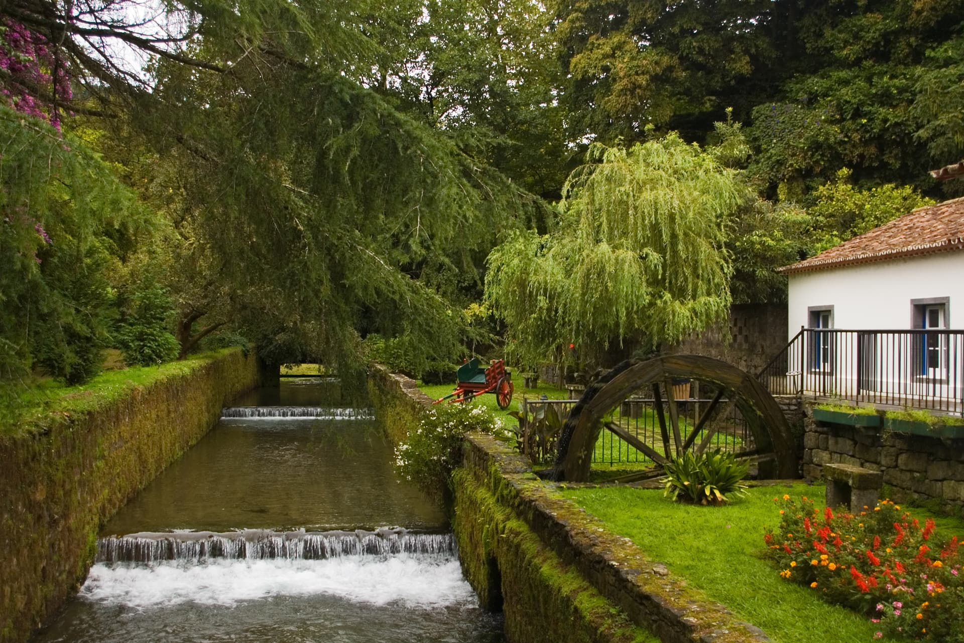 Water channel with small waterfalls, waterwheel, and white building in Furnas Town park.
